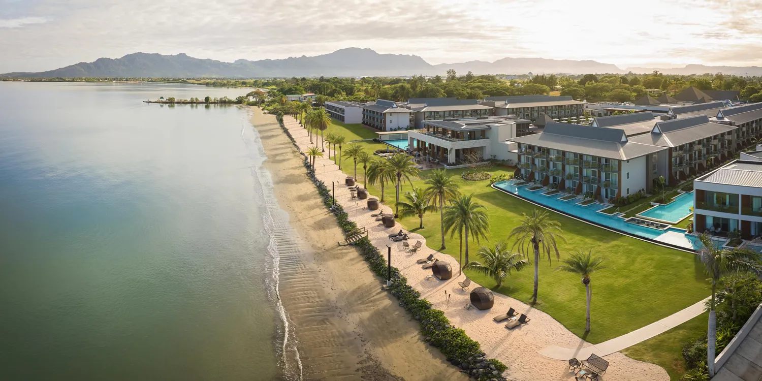 Panoramic view of the Crowne Plaza Fiji Nadi Bay Resort beachfront, with palm trees lining a sandy beach beside calm water and mountains in the distance.