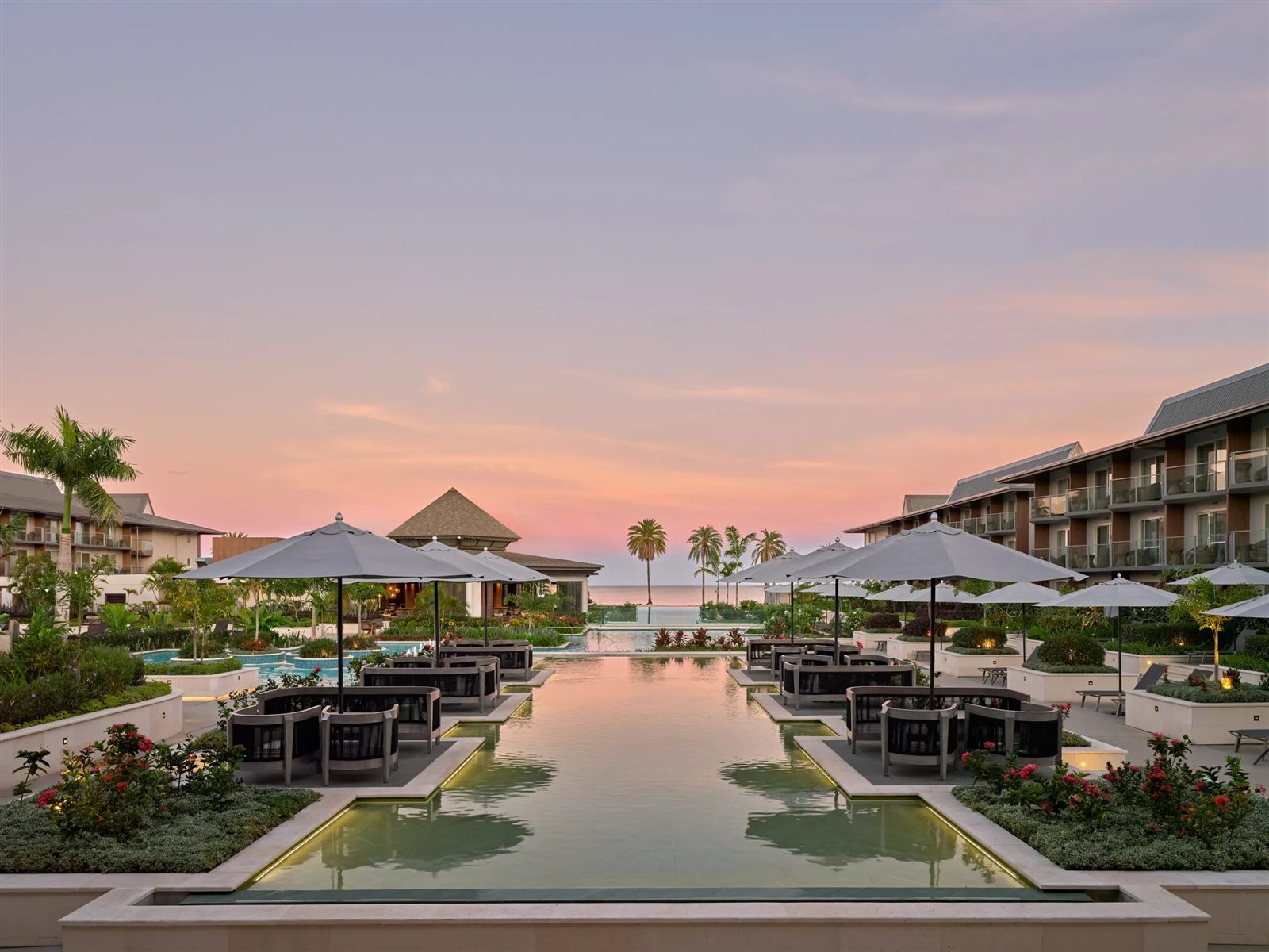 Resort pool terrace at sunset with umbrellas, lounge seating, and palm trees reflected in still water.